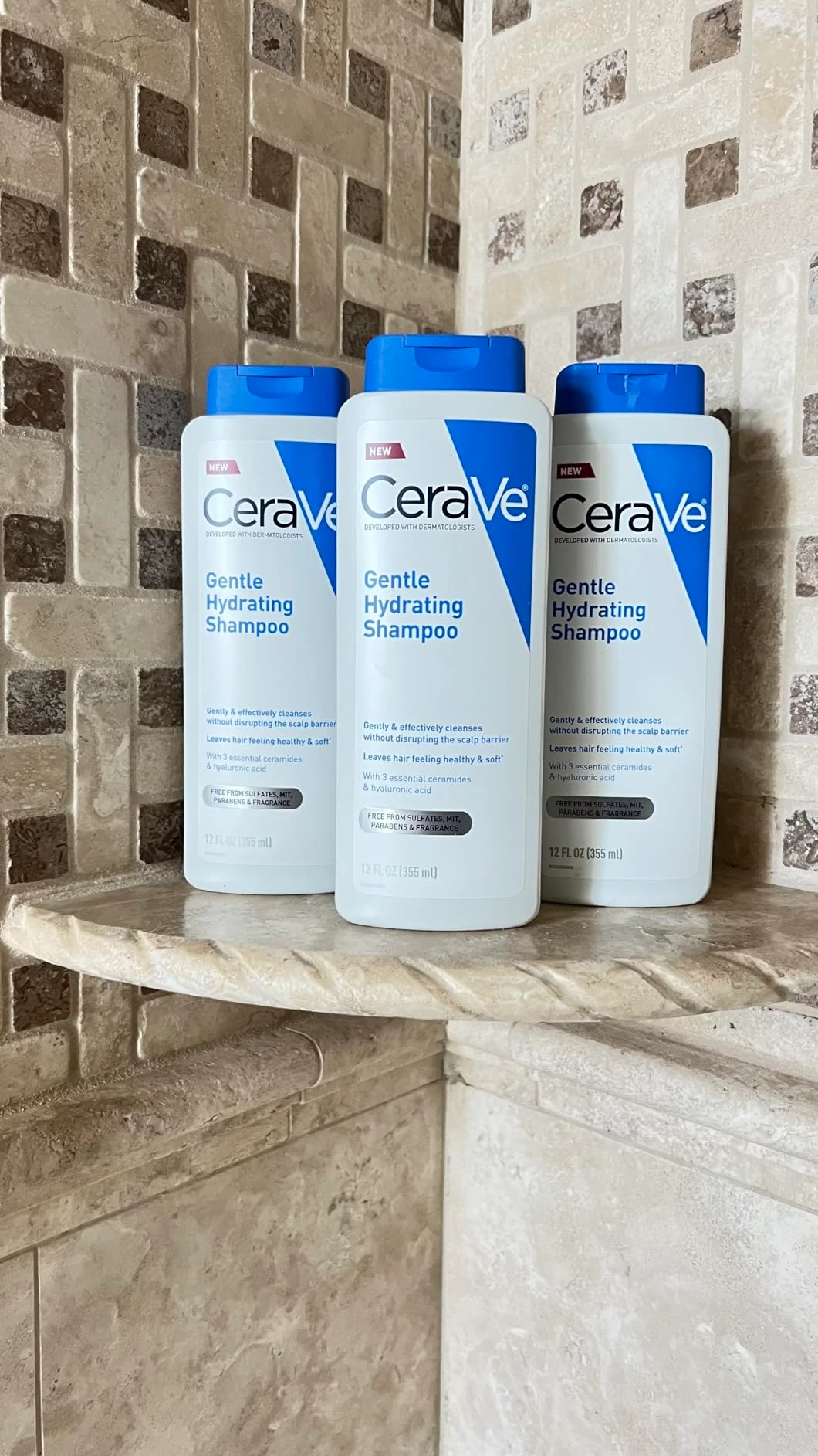 Ba chai dầu gội CeraVe Gentle Hydrating Shampoo màu trắng, nắp xanh dương, đặt trên kệ đá trong phòng tắm có tường gạch men màu be.