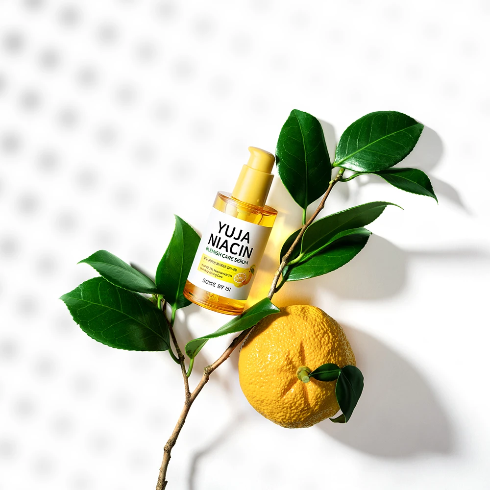 Chai serum Some By Mi Yuja Niacin Brightening Booster màu vàng, nằm trên cành cây có quả yuzu và lá xanh, nền trắng sáng
